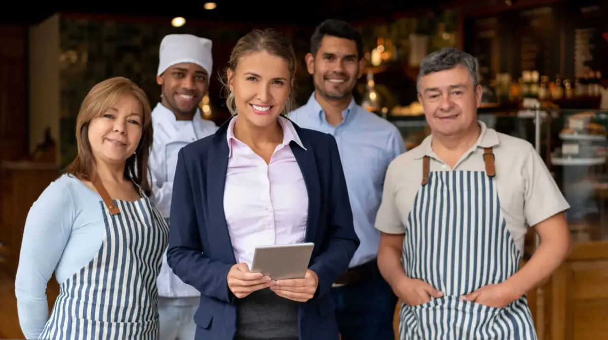 A hospitality manager holding a tablet stands with kitchen and front-of-house staff in a hotel dining area.