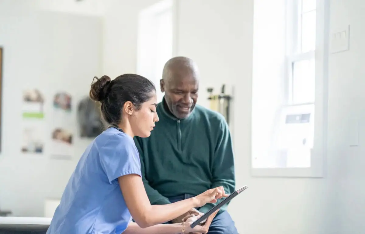 Home care nurse in blue scrubs reviewing information on a tablet with an older male client